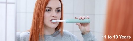 Banner photo of a young person with long red hair, looking in to a mirror using an electric toothbrush to clean their teeth.